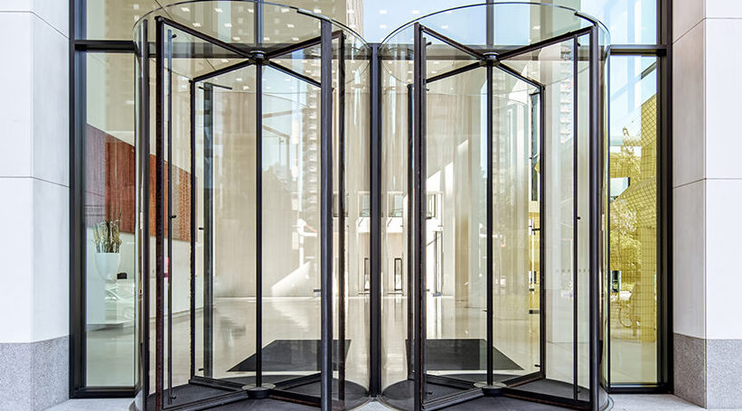Modern glass revolving door at office building entrance with reflections of cityscape, emphasizing sleek architectural design.